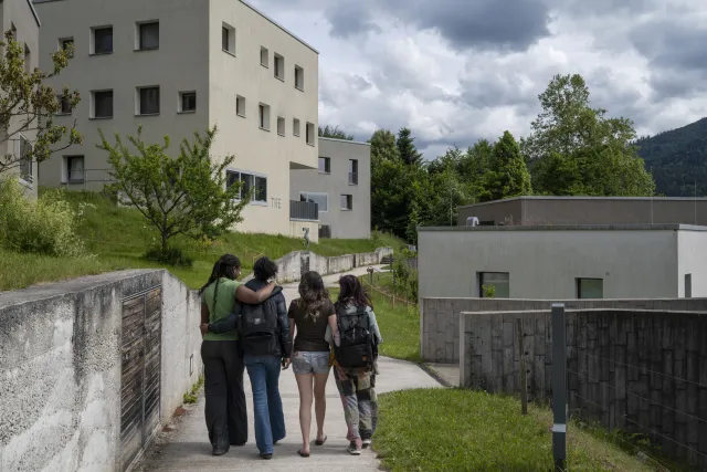 das Bild zeigt moderne, mehrstöckige Gebäude, in denen der Wohnbereich des UWC untergebracht ist. Die vier Freundinnen laufen Arm in Arm darauf zu.  